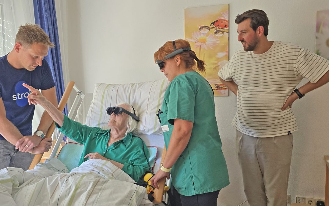 Zwei Mitarbeiter der Firma Stoll präsentieren eine VR-Brille für den Einsatz in der Ergotherapie. Eine Ergotherapeutin führt eine Therapie am Bett einer Bewohnerin durch. Dafür haben beide eine VR-Brille auf.